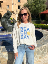 Woman wearing a 'Beach Day' sweatshirt standing in front of a fountain and building.