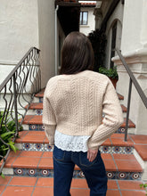 A woman viewed from behind wearing a cream cable-knit cropped sweater over a white lace top and blue jeans, standing on terracotta tiled stairs in front of a stucco building.