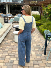 Woman in denim overalls standing on a patio with outdoor furniture and a building in the background.