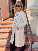 A woman wearing a long cream-colored coat over a striped top and dark pants, standing on outdoor terracotta steps with a stone wall and black metal railing in the background.