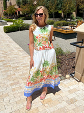 Woman wearing a floral top and skirt standing outdoors on a paved area with greenery.