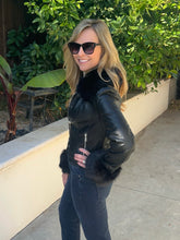 A woman wearing a black faux-leather moto jacket with black fur trim on the collar and cuffs, paired with dark gray jeans and black sunglasses, standing outdoors on a concrete walkway with green foliage in the background.