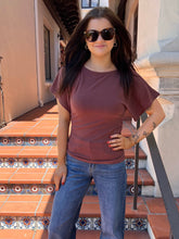 A woman wearing a short-sleeve crewneck t-shirt in a dusty rose-brown color, paired with blue jeans and sunglasses, standing on outdoor terracotta tile stairs.
