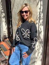 A woman wearing a dark charcoal gray knit sweater with large white floral patterns and blue jeans, standing outdoors against a textured stucco wall with leaf shadows.