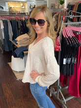 A woman wearing a cream-colored, long-sleeve lace-patterned top and blue jeans stands inside a clothing boutique, smiling and posing in front of racks of garments.