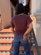 A person viewed from behind wearing a short-sleeve crewneck t-shirt in a dusty mauve-brown color and blue jeans, standing on outdoor tiled terracotta stairs.