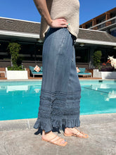 Person wearing a beige top and blue denim pants standing by a pool with a building and trees in the background.