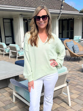 Woman in a light green sweater and white pants standing on a patio with outdoor furniture.
