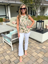 Woman standing outdoors on a patio wearing a patterned top and white pants.