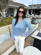 Woman in a blue top and white pants standing outdoors with a building and patio furniture in the background.
