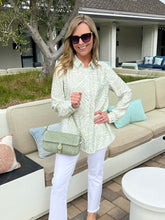 Woman in a light green patterned shirt and white pants standing in an outdoor setting.