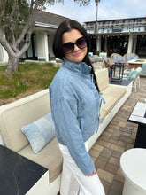 Woman wearing a blue jacket and sunglasses in an outdoor setting with a building and trees in the background.
