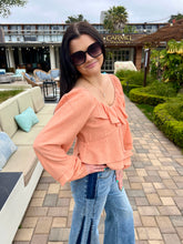 Woman in peach blouse and blue jeans standing outdoors with a building and palm trees in the background