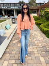 Woman in peach blouse and blue jeans standing on a patio with outdoor furniture and greenery.