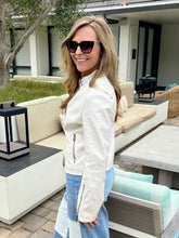 Woman wearing a white jacket and sunglasses outdoors in a modern outdoor setting.