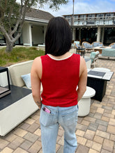 Person wearing a red tank top and light blue jeans standing in an outdoor setting with furniture and a building in the background.