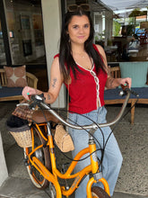 Woman in a red top and blue jeans standing next to a yellow bicycle with a basket in an indoor setting.