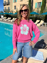 Woman in pink sweater with a white floral design standing by a pool.
