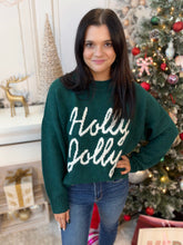 A woman wearing a dark green knit sweater with 'Holly Jolly' written in white script across the front, standing in a decorated holiday room next to a Christmas tree.