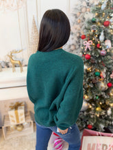 A woman with dark hair is shown from behind wearing a dark green knit sweater and blue jeans, standing in a decorated indoor setting with a Christmas tree and holiday decorations.