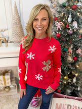 A smiling woman wearing a bright red crewneck sweater with white and gold snowflake patterns, paired with blue jeans, standing in front of a decorated Christmas tree and holiday decor.