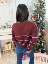 A woman viewed from behind wearing a ribbed knit sweater with horizontal stripes in burgundy and dusty pink, paired with blue jeans, standing in a decorated holiday room next to a Christmas tree.