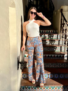 Women posing on stairs with hand on sunglasses, colorful pants and white tank top.